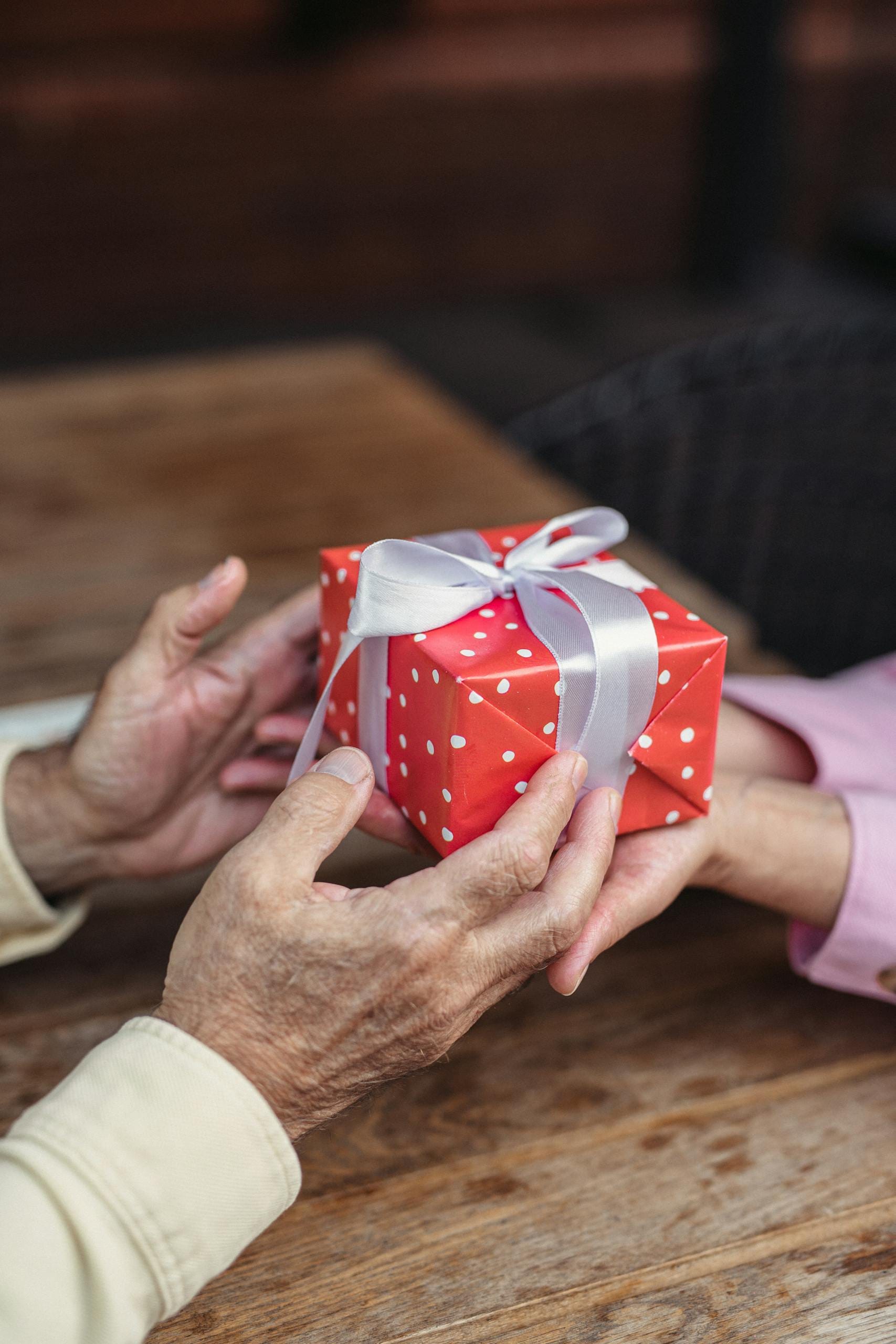 Close-up of elderly hands exchanging a red gift box with a white ribbon on a wooden table.